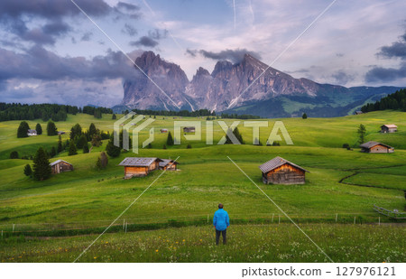 Hiker standing in a green meadow with wooden huts in mountains 127976121