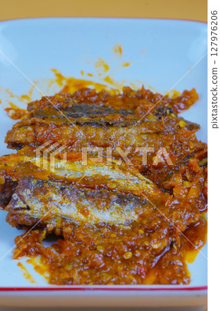 A rectangular white plate holds several pieces of fried mackarel tuna fish in sambal balado or a rich, spicy chilli sauce, set against a plain yellow background. 127976206