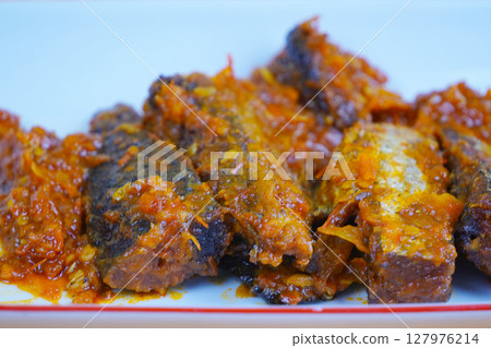 A rectangular white plate holds several pieces of fried mackarel tuna fish in sambal balado or a rich, spicy chilli sauce, set against a plain yellow background. 127976214