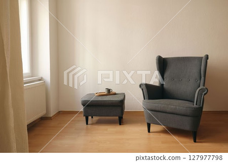 Cozy reading corner. Dark grey armchair, matching ottoman in minimalist living room near window. Cup of tea, coffee. Natural light, empty copy space. Elegant Scandinavian interior. Beige wall Cozy reading corner. Dark grey armchair, matching ottoman in minimalist living room near window. Cup of tea, coffee. Natural light, empty copy space. Elegant Scandinavian interior. Beige wall 127977798