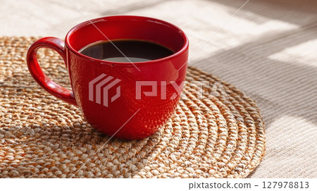 A vibrant red cup of dark coffee sits on a woven mat, bathed in soft sunlight, offering a moment of simple pleasure and warmth. A vibrant red cup of dark coffee sits on a woven mat, bathed in soft sunlight, offering a moment of simple pleasure and warmth. 127978813