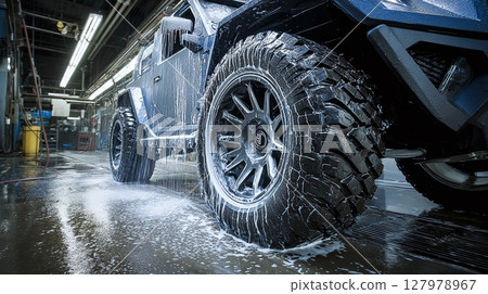 Vehicle is getting a cleaning with soap and water. The soap runs off the tires and frame. The car is black and has big wheels and tires. 127978967