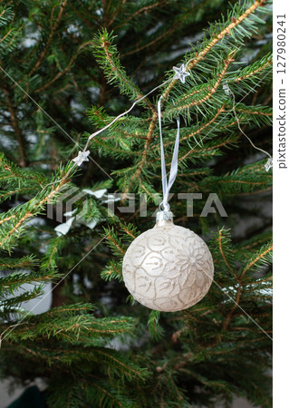 Close-up of a delicate Christmas ornament resting on a lush tree branch, showcasing intricate details and festive brilliance in warm holiday hues Close-up of a delicate Christmas ornament resting on a lush tree branch, showcasing intricate details and festive brilliance in warm holiday hues 127980241