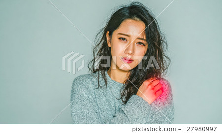 An Asian woman experiencing shoulder pain, gently touching her shoulder with a visible red area indicating discomfort An Asian woman experiencing shoulder pain, gently touching her shoulder with a visible red area indicating discomfort 127980997