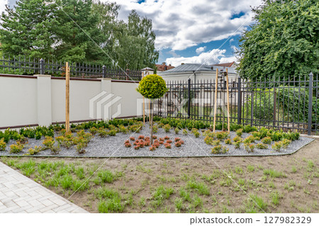 Back and front yard cottage garden, flowering plant and green grass lawn, brown pavement and orange brick wall, evergreen trees on background, in good care maintenance landscaping in park 127982329