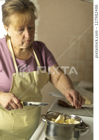A senior woman puts pieces of sweet butter into a saucepan. A senior woman puts pieces of sweet butter into a saucepan. 127982486