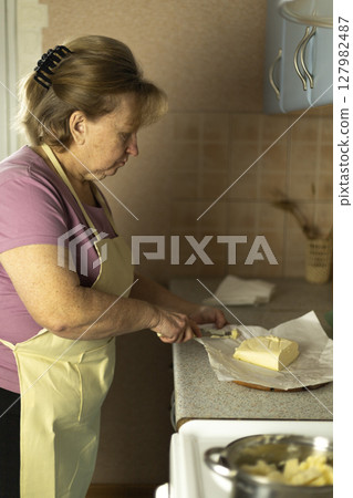 A senior woman is adding sweet cream butter to a saucepan on the stove. A senior woman is adding sweet cream butter to a saucepan on the stove. 127982487