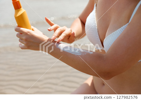Hand of a young woman applying sun cream or sunscreen on her tanned arm to protect her skin from the sun. Portrait of woman holding suntan lotion or  suntan moisturizer. 127982576
