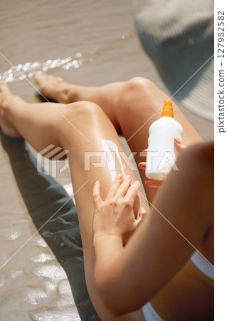 Woman applying sunscreen to her legs while lounging on the beach during a sunny day, focusing on skincare and sun protection. Skincare concept 127982582