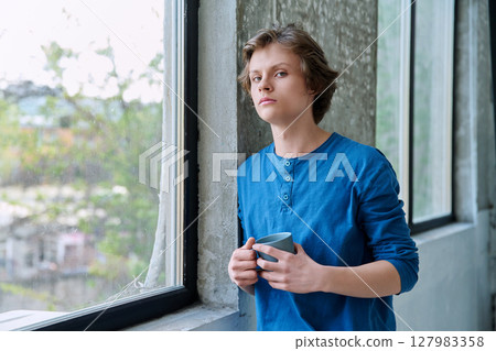 Teenage serious guy with cup in hands in home near window, looking at camera 127983358