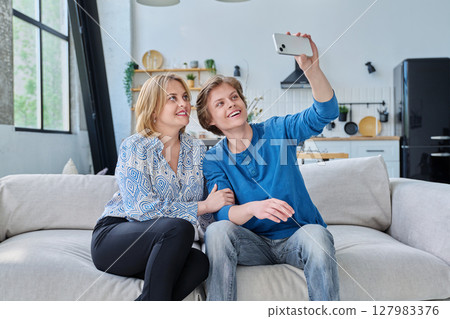 Middle aged mother and teenage son sitting together at home taking selfie photo on smartphone 127983376