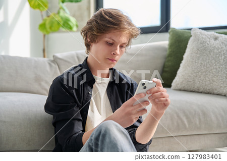 Serious handsome teenager guy using smartphone sitting on floor at home. 127983401