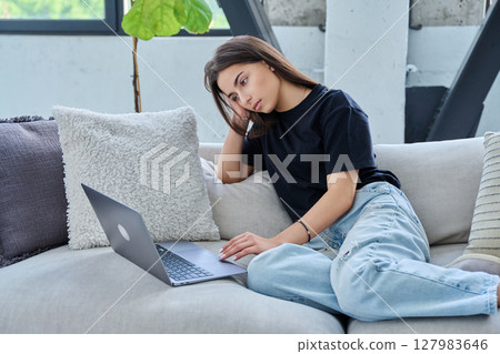 Serious teenage girl looking at laptop, sitting on sofa at home Serious teenage girl looking at laptop, sitting on sofa at home 127983646