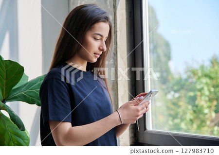 Teenage girl using smartphone standing at home near window, copy space Teenage girl using smartphone standing at home near window, copy space 127983710