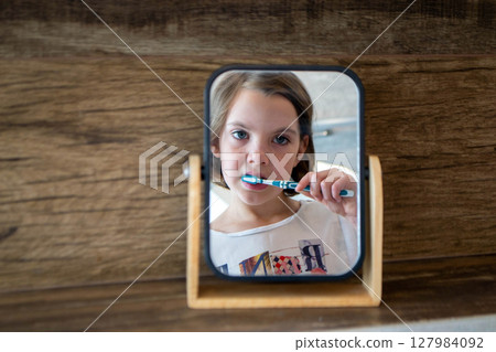 Oral hygiene, healthy teeth and care. Girl brushing teeth with toothbrush and looking in mirror in bathroom interior in the morning, closeup, empty space. High quality photo Oral hygiene, healthy teeth and care. Girl brushing teeth with toothbrush and looking in mirror in bathroom interior in the morning, closeup, empty space. High quality photo 127984092