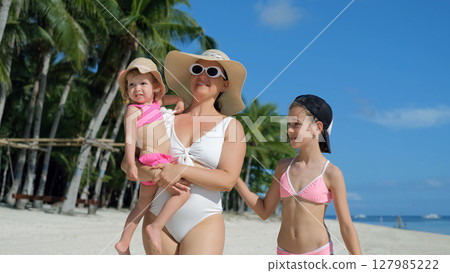 Mom walks on tropical beach shoreline with baby and older daughter 127985222