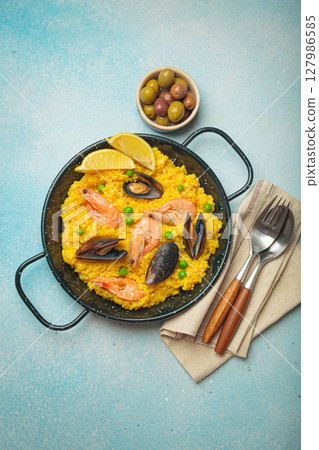 Spanish seafood Paella with shrimps, mussels, peas and lemon in a pan on light blue background top view. Mediterranean meal Spanish seafood Paella with shrimps, mussels, peas and lemon in a pan on light blue background top view. Mediterranean meal 127986585