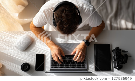 Professional videographer working from home, typing on laptop with headphones, surrounded by camera gear, natural light filling workspace 127986949