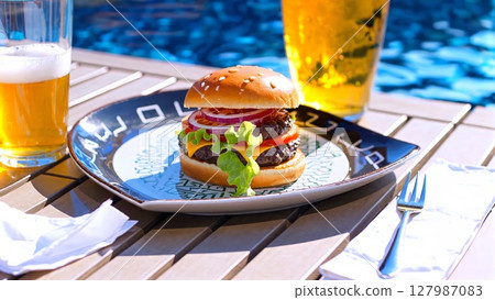 Poolside hamburger featuring fresh ingredients, golden beer glasses, relaxing summer dining experience with grilled beef patty and crisp vegetable toppings 127987083