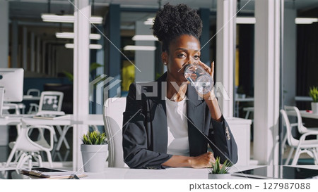 Professional African American businesswoman sipping bottled water while seated at workspace, pausing during workday and maintaining hydration at modern office desk Professional African American businesswoman sipping bottled water while seated at workspace, pausing during workday and maintaining hydration at modern office desk 127987088