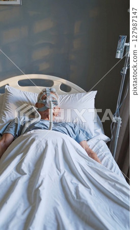 Elderly patient wearing respiratory mask lying in hospital bed, undergoing treatment for sleep apnea or respiratory illness, demonstrating healthcare and recovery 127987147