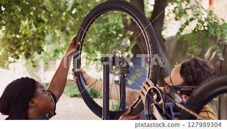 African american girlfriend carrying wheel to reattach on bicycle body with caucasian boyfriend. Sports-loving multiethnic couple carefully securing dismantled bike tire outside. 127989304