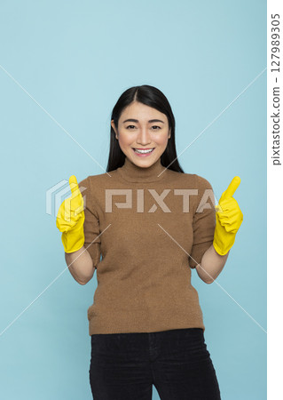 Happy asian housekeeper posing in blue background studio, expressing satisfaction with household chores. Young female janitor gesturing two thumbs up during productive morning cleanup routine. 127989305