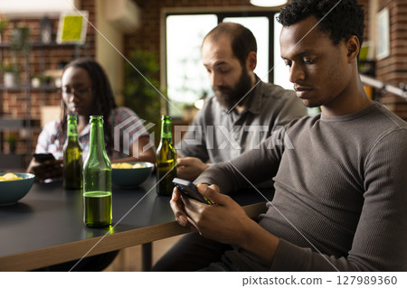 Close up of african american man using smartphone, with bottle of beer nearby on table. Black male individual engages in online browsing while sitting with friends in brick walled room. 127989360