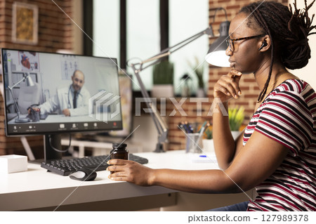 Self employed woman speaking with doctor on video call, discussing diagnosis and side effects while holding prescription medication. Remote female patient consults physician from home. Self employed woman speaking with doctor on video call, discussing diagnosis and side effects while holding prescription medication. Remote female patient consults physician from home. 127989378