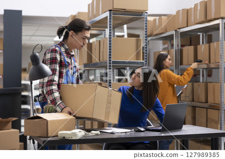 Asian workers team handles business logistics in a compact inventory room, taping boxes and attaching shipping labels. Small scale warehouse staff ensures retail product delivery. 127989385