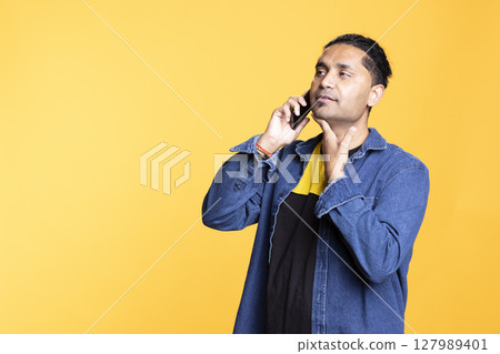 Indian person smiling during a conversation with his group of friends, chatting on telephone line against an orange background. Young adult with a man bun in casual attire gossiping. 127989401