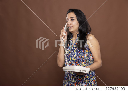 Female model talking on landline, having conversation with family or friends. Indian woman on a call, holding telephone receiver to her ear and standing against brown background. 127989403