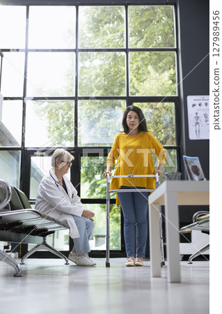 Asian woman using walking frame during rehabilitation healing process, receiving physical support from medical specialist at a health clinic. Moving around for recovery journey. Asian woman using walking frame during rehabilitation healing process, receiving physical support from medical specialist at a health clinic. Moving around for recovery journey. 127989456