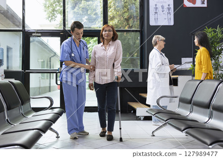 Friendly nurse helps an asian patient with mobility disability walk on crutches through a hospital lobby, highlighting rehabilitation, support and inclusive healthcare services. Healing process. Friendly nurse helps an asian patient with mobility disability walk on crutches through a hospital lobby, highlighting rehabilitation, support and inclusive healthcare services. Healing process. 127989477
