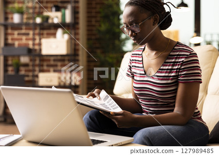 Determined female entrepreneur carefully reviewing printed documents during remote work session. Focused black woman flips through clipboard, preparing notes for upcoming project presentation. 127989485