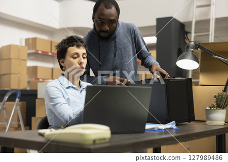 Diverse employees at small business fulfillment center manage local shipping and inventory processes, working together in a warehouse full of taped boxes and storage shelving units. 127989486