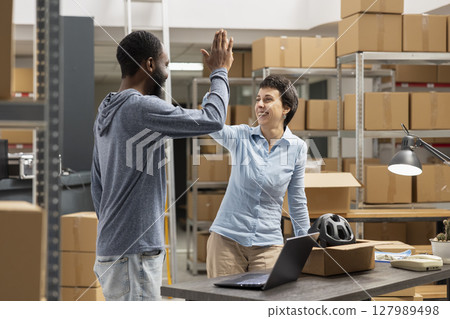 Two small business workers celebrating progress with a high five, symbolizing teamwork and achievement in e commerce startup. People working in supply room packed with boxes and goods. 127989498