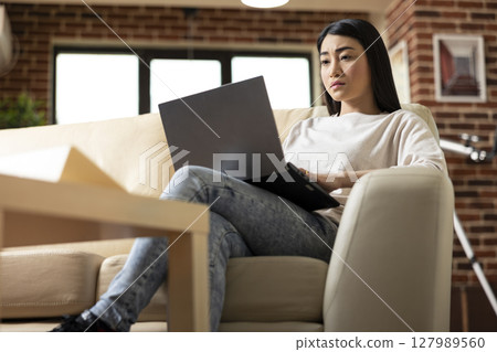 Serious female manager working remotely in brick wall apartment, typing on laptop and preparing business reports. Self employed asian woman showing productivity at home, reviewing company proposals 127989560