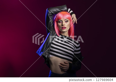 Strong elegant woman projects attitude in studio using bold expression and confident posture. Fierce female model with pink hair, wearing black leather jacket poses against isolated background. Strong elegant woman projects attitude in studio using bold expression and confident posture. Fierce female model with pink hair, wearing black leather jacket poses against isolated background. 127989604