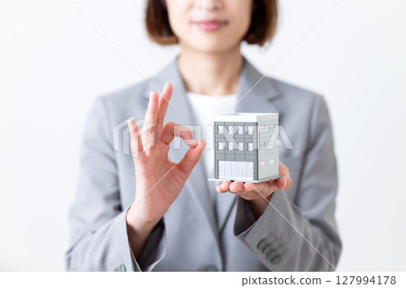 一位身穿灰色西裝的中年婦女拿著房屋模型，做著圓圈手勢 127994178