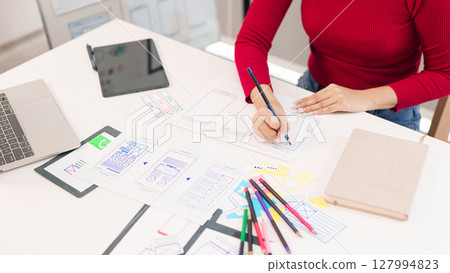 Asian Woman UI UX Designer Creating Mobile App Interface on Desk with Laptop Tablet and Sketches User Experience Design Process and Hand 127994823
