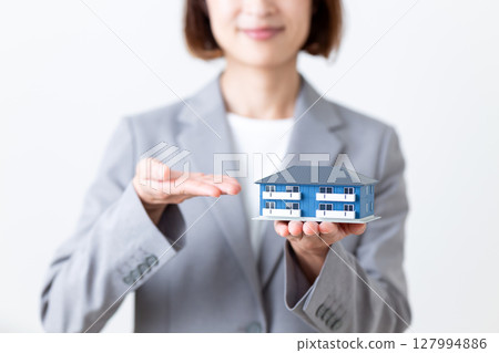 A middle-aged woman in a gray suit holding a model of an apartment house and recommending it 127994886