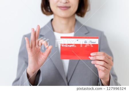A middle-aged woman in a gray suit holding a bank deposit book and making a circle sign 127996324