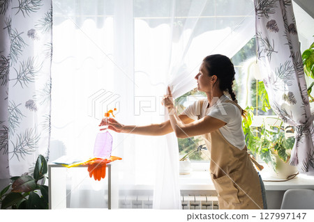 Woman in rubber gloves and apron manually washes window of house with rag cleaner and mop inside interior with home plants on windowsill. Restoring order and cleanliness in spring, cleaning servise Woman in rubber gloves and apron manually washes window of house with rag cleaner and mop inside interior with home plants on windowsill. Restoring order and cleanliness in spring, cleaning servise 127997147