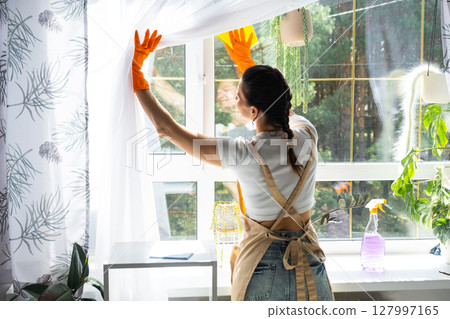 Woman in rubber gloves and apron manually washes window of house with rag cleaner and mop inside interior with home plants on windowsill. Restoring order and cleanliness in spring, cleaning servise 127997165