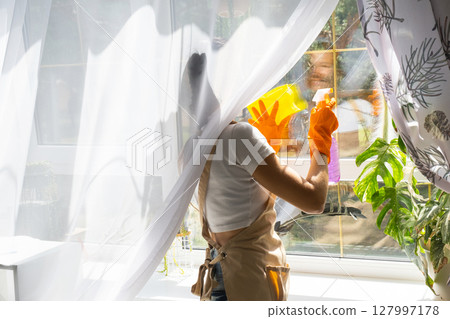 Woman in rubber gloves and apron manually washes window of house with rag cleaner and mop inside interior with home plants on windowsill. Restoring order and cleanliness in spring, cleaning servise 127997178