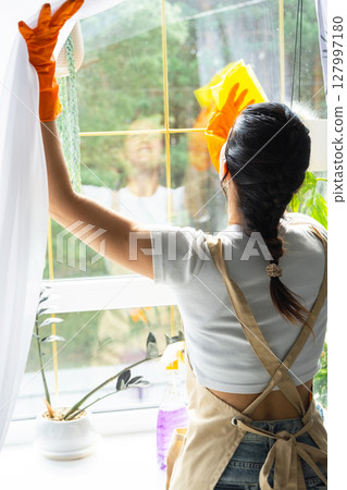 Woman in rubber gloves and apron manually washes window of house with rag cleaner and mop inside interior with home plants on windowsill. Restoring order and cleanliness in spring, cleaning servise Woman in rubber gloves and apron manually washes window of house with rag cleaner and mop inside interior with home plants on windowsill. Restoring order and cleanliness in spring, cleaning servise 127997180