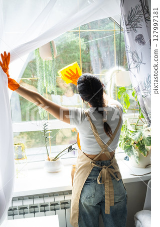 Woman in rubber gloves and apron manually washes window of house with rag cleaner and mop inside interior with home plants on windowsill. Restoring order and cleanliness in spring, cleaning servise 127997181