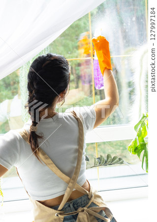 Woman in rubber gloves and apron manually washes window of house with rag cleaner and mop inside interior with home plants on windowsill. Restoring order and cleanliness in spring, cleaning servise 127997184