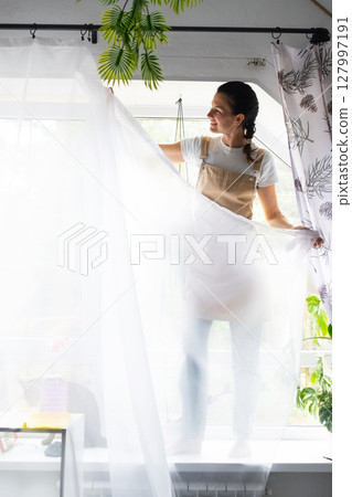 Woman in apron hangs transparent tulle curtains on large attic windows in the house inside the interior with potted plants. Spring cleaning, tidying up 127997191